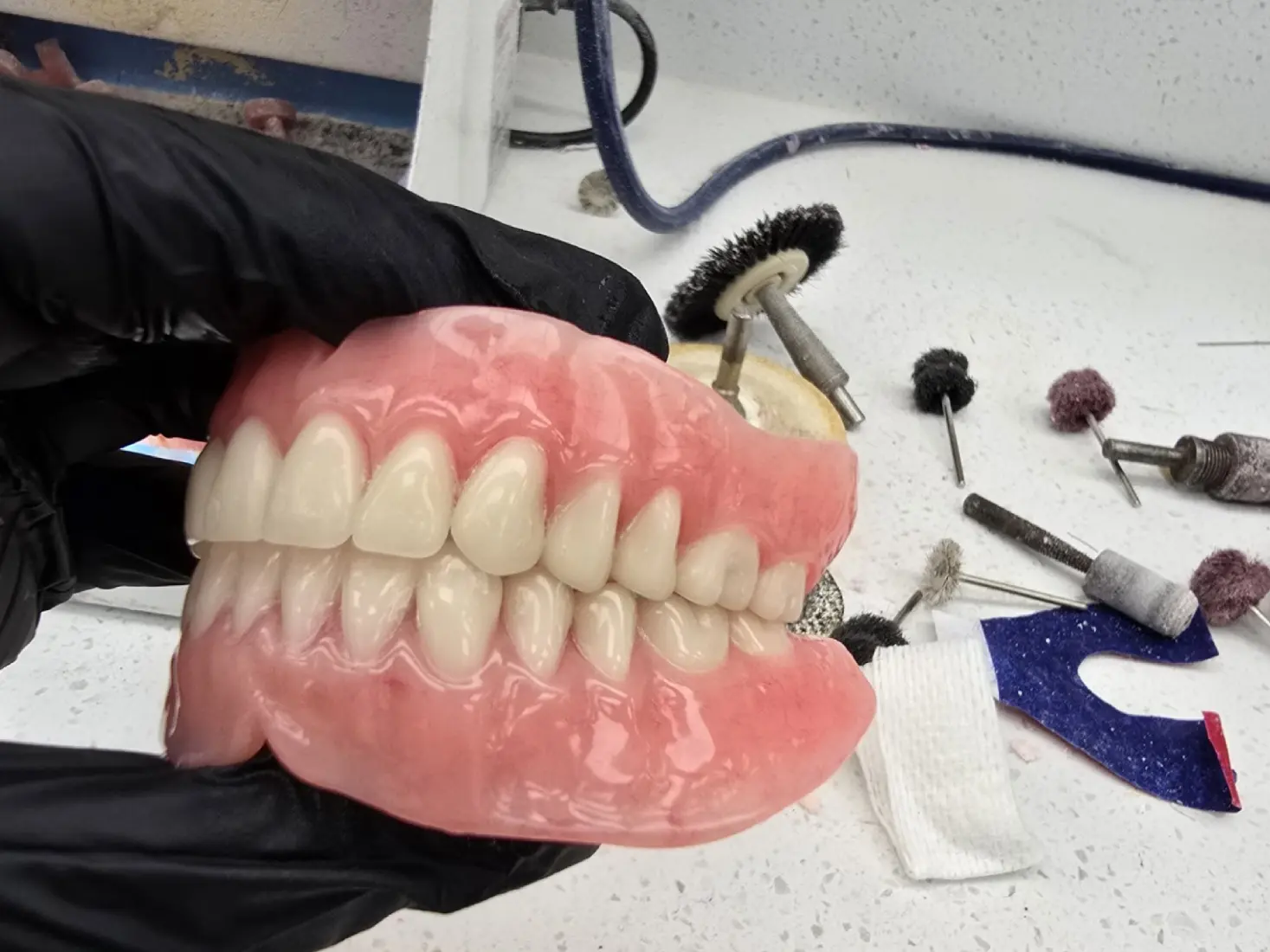2 A hand wearing a black glove holds a set of complete dentures, with dental tools and materials on a white countertop in the background.
