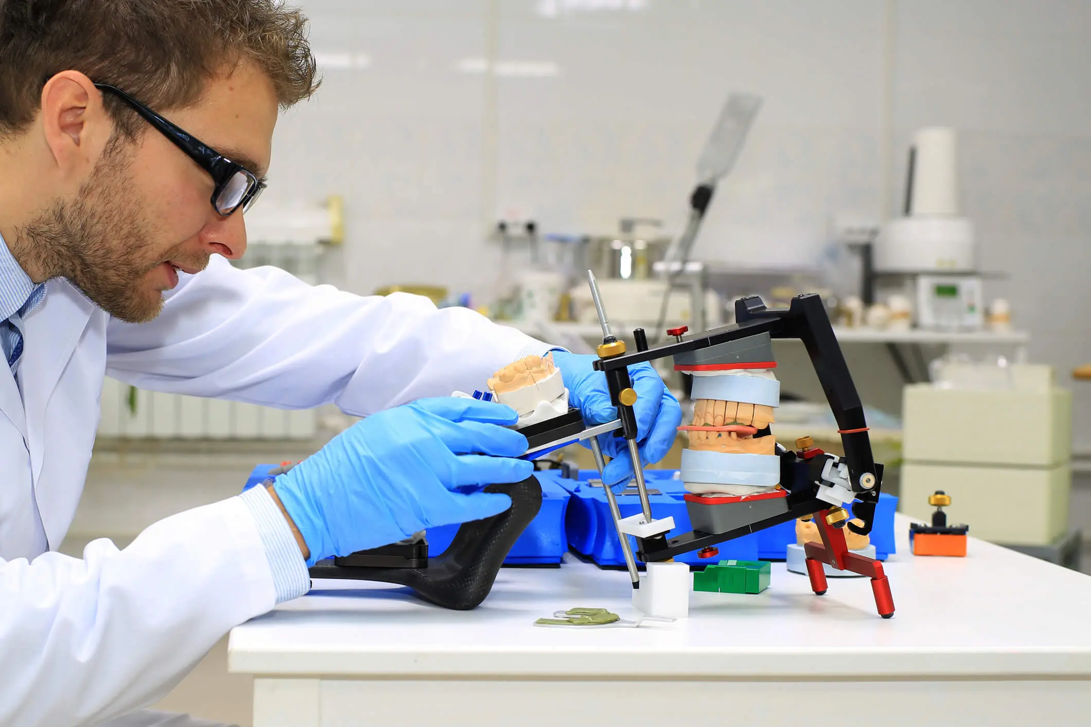 GettyImages-618843434 A man in a lab coat and gloves adjusts a dental articulator with dental models in a laboratory setting.